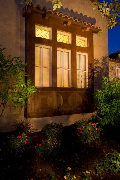Warmly lit window with decorative woodwork, surrounded by colorful flowers at dusk.